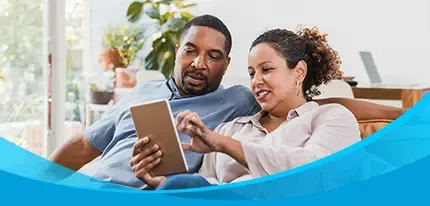 A couple relaxes on a couch together browsing a tablet in a bright living room.